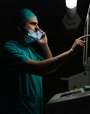 A clinician in surgical scrubs and mask making a phone call while reading a patient record on a lit screen in a darkened treatment room.